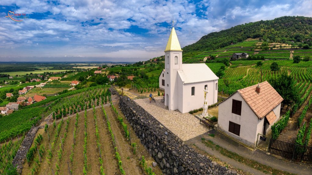 A scenic aerial view of a white church with a golden steeple, surrounded by lush vineyards and rolling hills in Hungary’s wine region.