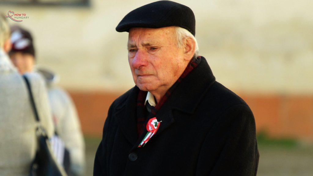 Elderly Hungarian man in a black coat and hat wearing a kokárda on March 15th, standing with a solemn expression during the National Day commemorations