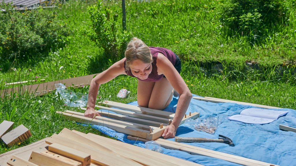 Woman assembling IKEA furniture outdoors on a blue blanket in a grassy garden, surrounded by wooden parts, tools, and an instruction manual.