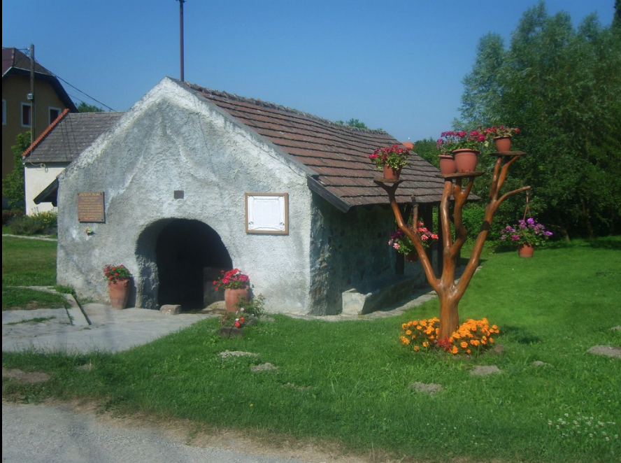 Historic stone washhouse and town well in a Hungarian village, built in the 1700s, featuring a tiled roof, arched entrance, flower pots, and decorative landscaping.