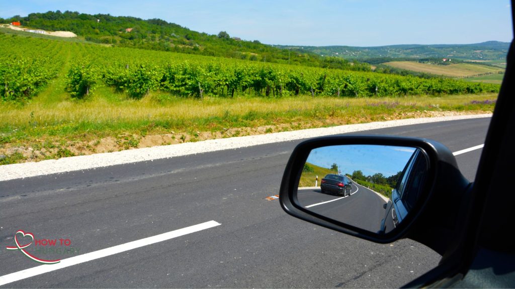 A car driving on a Hungarian motorway with a scenic view visible in the side mirror, showing countryside or road landscape