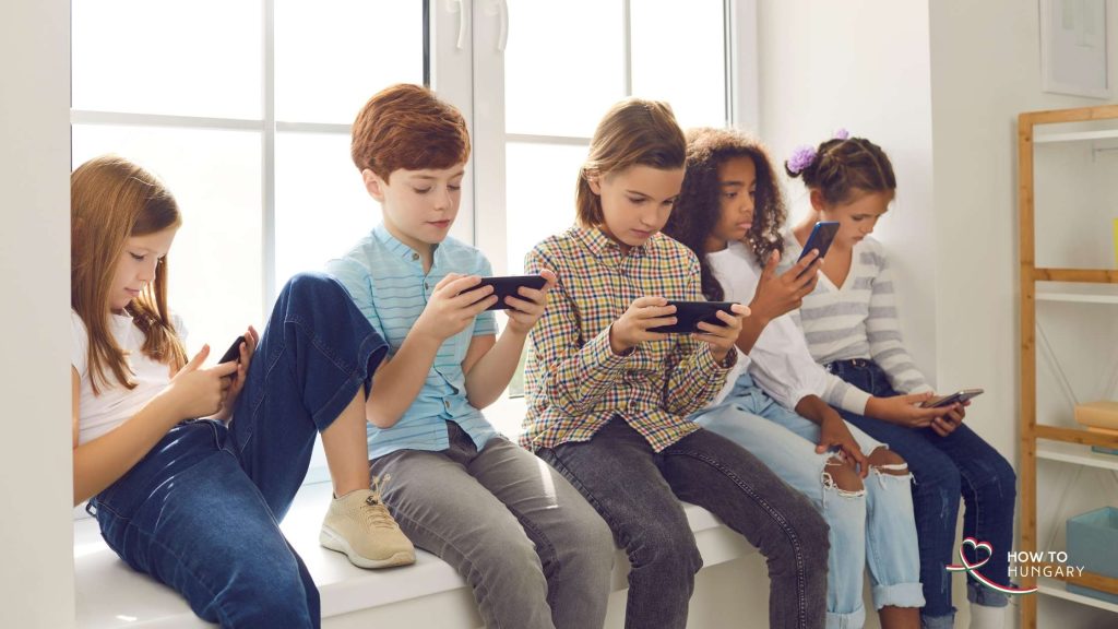 Five children sitting on a windowsill glued to their phones, illustrating phones banned in Hungarian schools in 2026