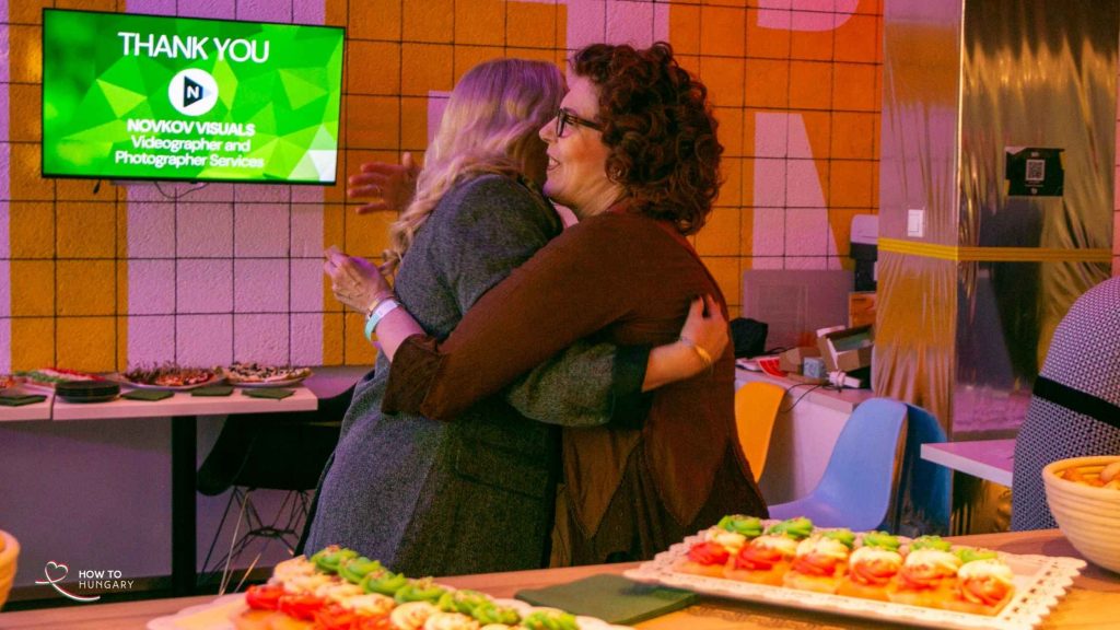 A hug by the food table as a NOVKOV Visuals thank-you shows on screen at the HOW TO HUNGARY September Social, KLUSTER Budapest.